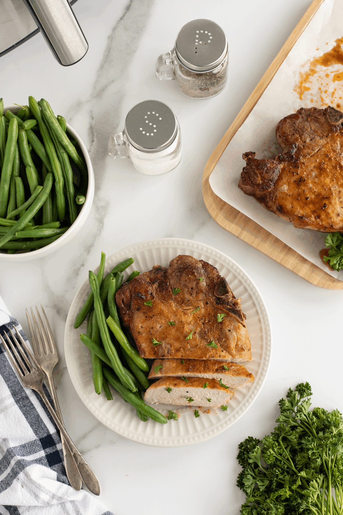A plate with a cooked pork chop, some slices cut, garnished with parsley, and green beans. Nearby are a bowl of green beans, salt and pepper shakers, parsley, and a tray with another pork chop. Forks and a napkin are beside the plate.