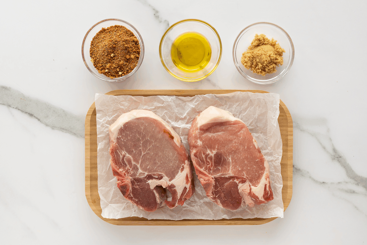 Two raw pork chops on parchment paper on a wooden board, with small bowls of brown sugar, olive oil, and dry seasoning arranged above them on a white marble surface.