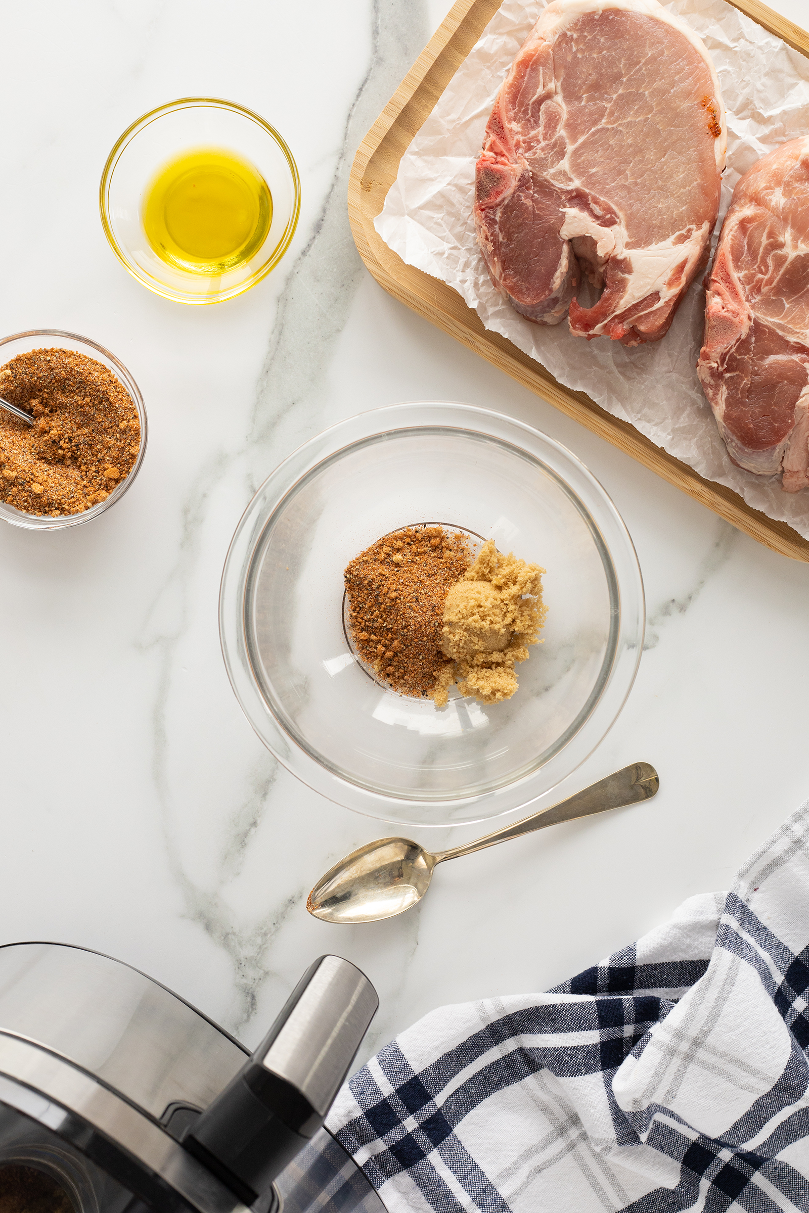 A glass bowl with brown sugar and spices, a spoon, a small bowl of oil, a dish of spice mix, two raw pork chops on a wooden tray, and a striped towel on a white marble countertop.