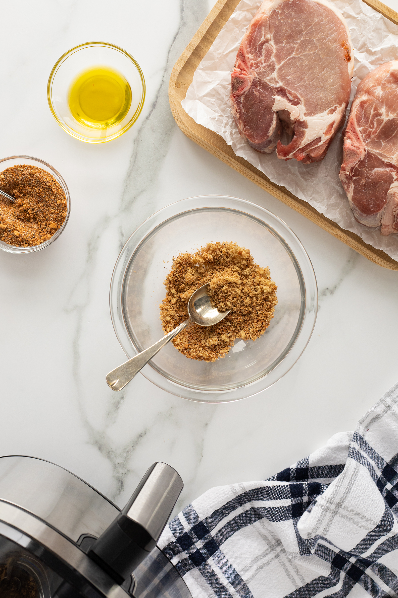 Overhead view of raw pork chops on a tray, a bowl of olive oil, a bowl of spice rub with a spoon, and a kitchen towel on a marble countertop. Part of an air fryer is visible in the corner.