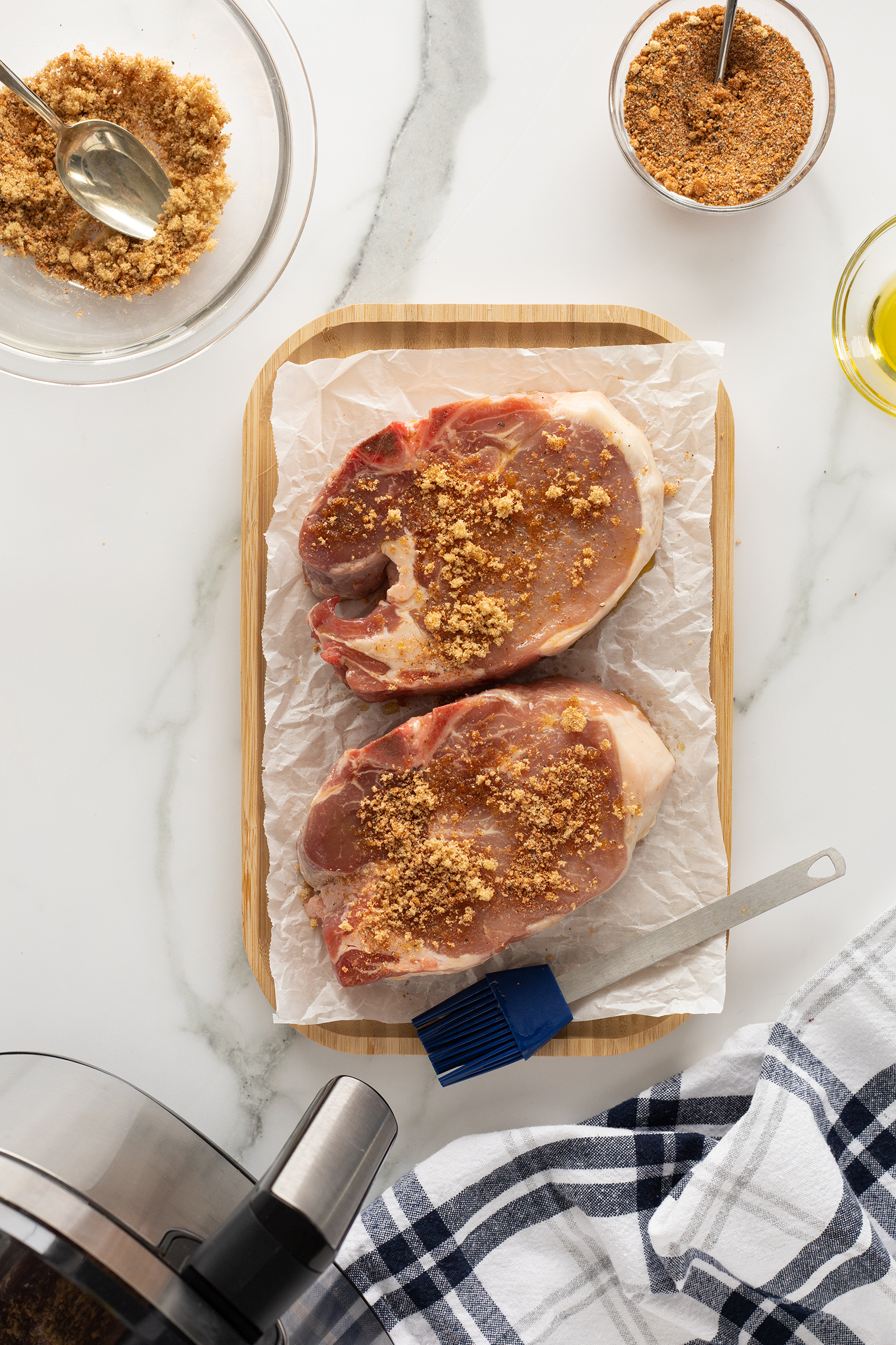 Two raw pork chops on parchment paper atop a wooden board, sprinkled with brown sugar seasoning. A basting brush, bowl of seasoning, oil, and checkered towel are nearby on a marble surface.