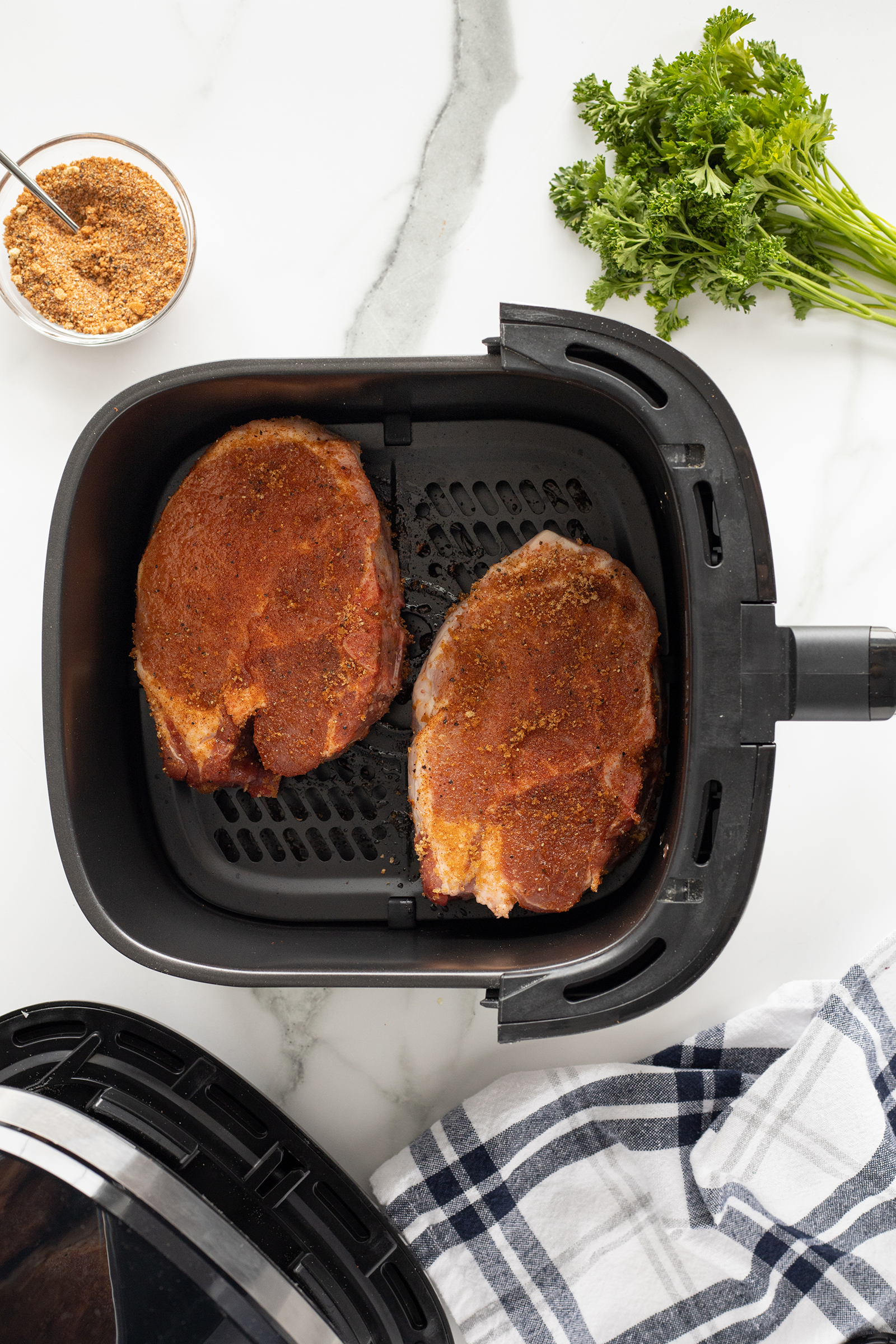 Two seasoned pork chops sit in an open air fryer basket on a marble counter, with a small bowl of spice mix, a bunch of fresh parsley, and a striped cloth nearby.