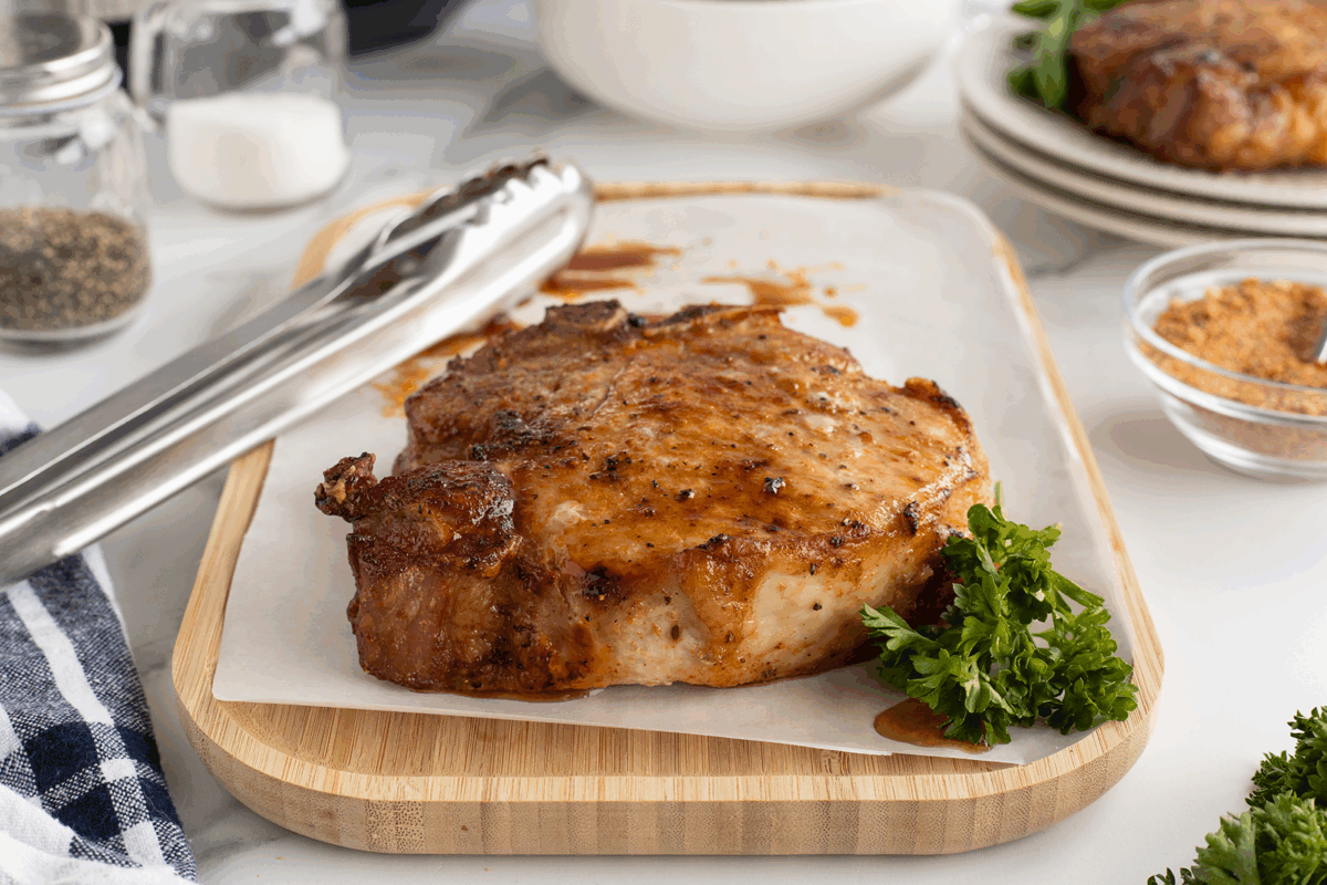 A cooked, seasoned pork chop rests on parchment paper atop a wooden cutting board with metal tongs beside it. Fresh parsley garnishes the chop, and various spices and plates appear in the background.
