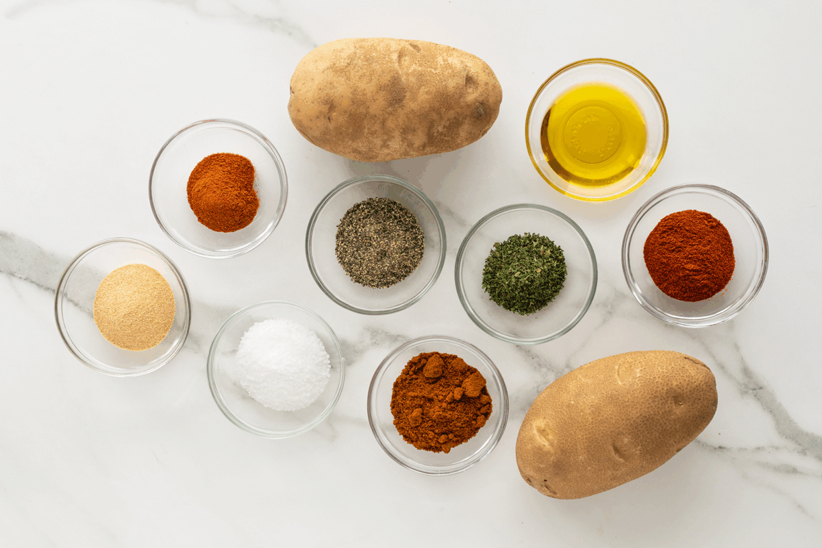 Two whole russet potatoes and small glass bowls filled with olive oil, black pepper, parsley, salt, garlic powder, paprika, smoked paprika, and chili powder are arranged on a white marble surface.