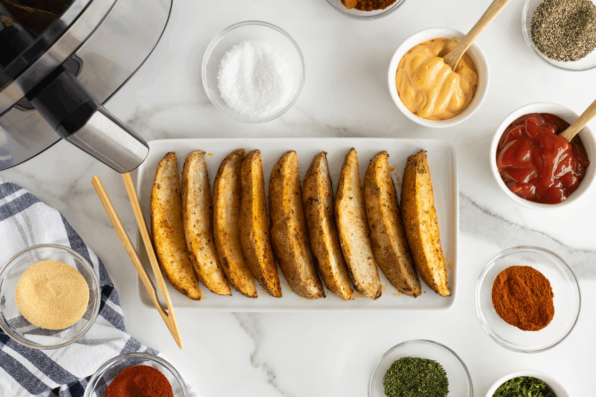 A rectangular plate with seasoned potato wedges is surrounded by bowls of spices, herbs, salt, ketchup, mayonnaise, and an air fryer on a white marble surface. Wooden tongs rest on the plate.