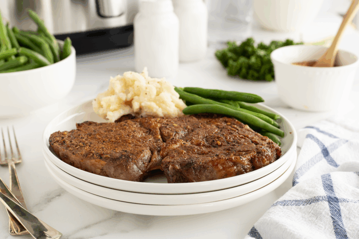 A plate with two seasoned steaks, mashed potatoes, and green beans on a white table. Nearby are a bowl of green beans, salt and pepper shakers, parsley, a sauce bowl, and a striped napkin.