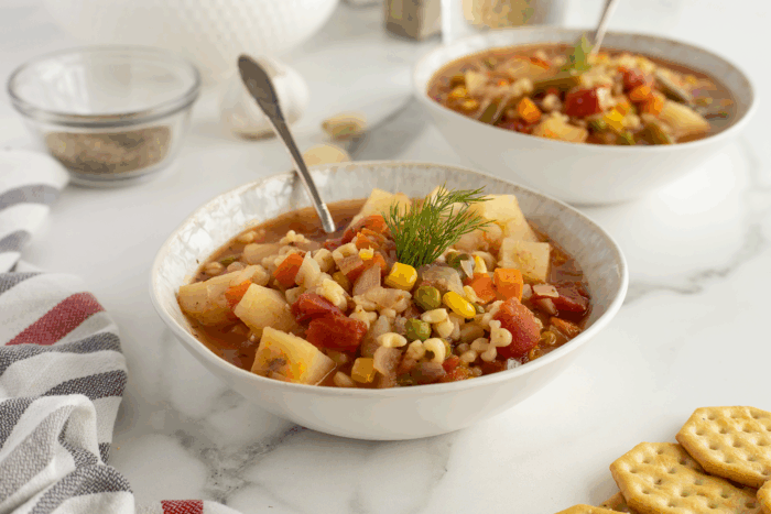 Two bowls of colorful vegetable soup with pasta and fresh dill garnish sit on a white marble surface. Spoons rest in each bowl, with crackers and a striped napkin nearby. The background includes seasonings and garlic.