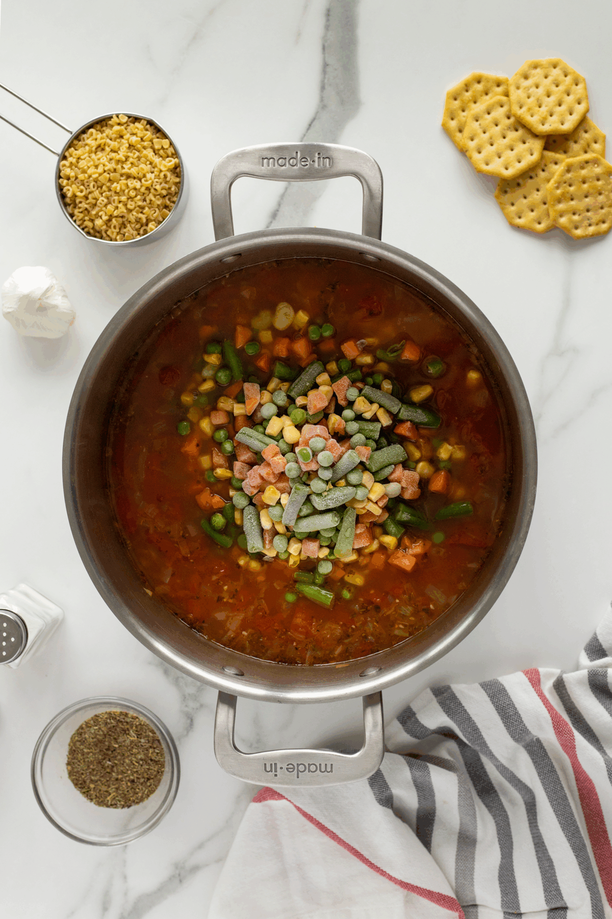 A pot of vegetable soup with frozen mixed vegetables on top sits on a marble countertop, surrounded by small pasta, round crackers, garlic, a bowl of herbs, and a striped kitchen towel.