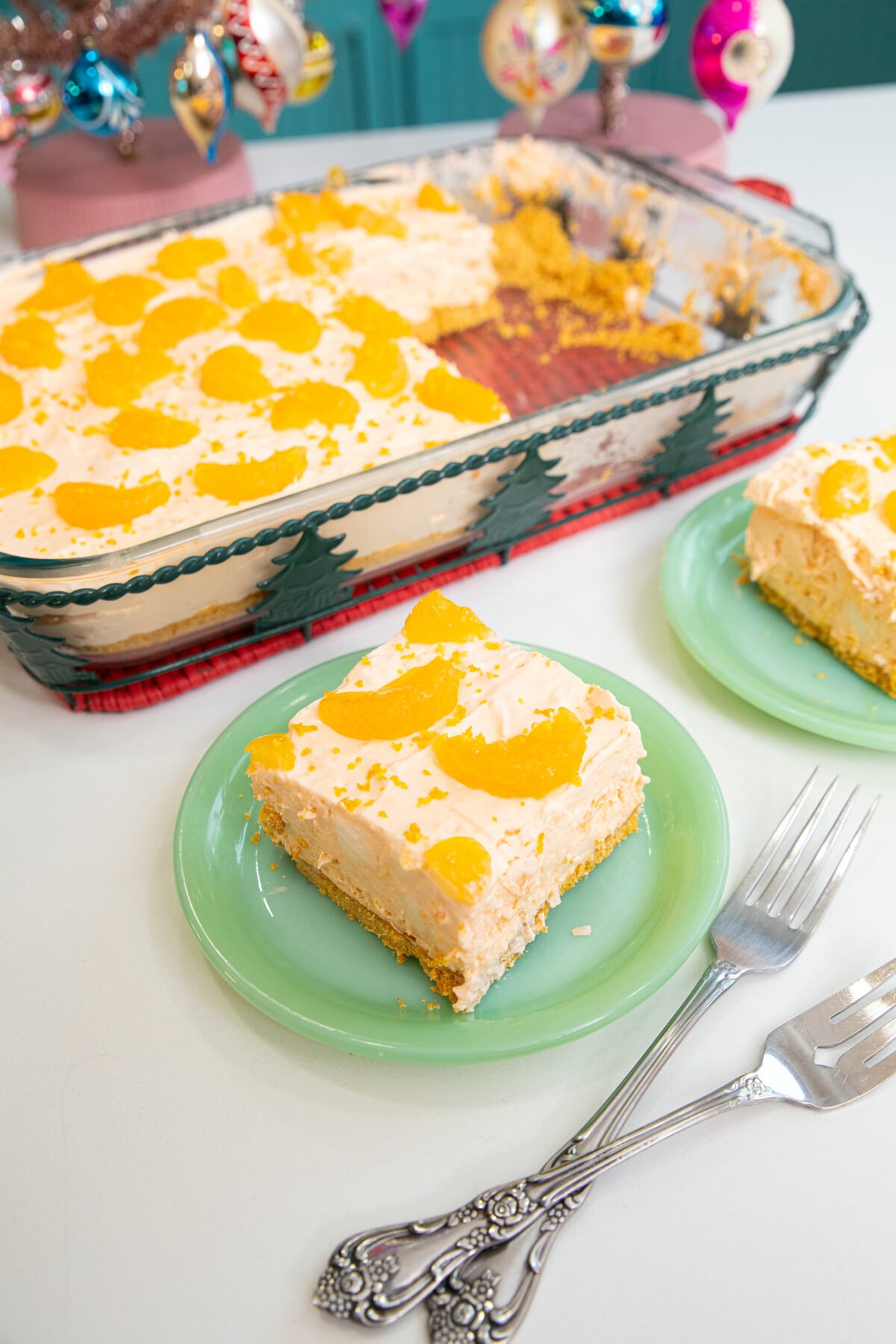 A glass baking dish of creamy dessert topped with mandarin orange slices sits on a festive tray. Two servings are plated on green dishes with vintage silver forks beside them. Ornaments decorate the background.