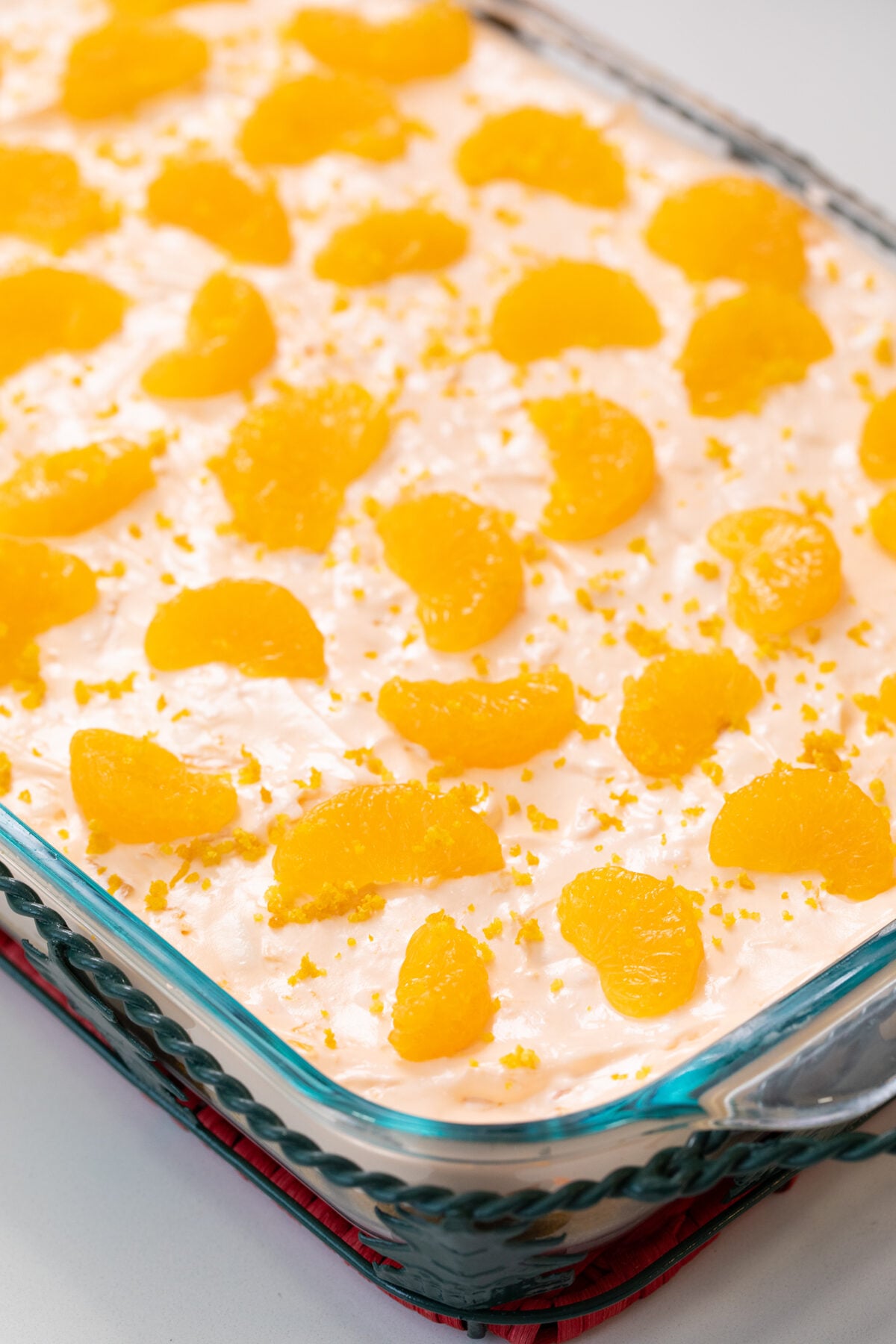 A glass baking dish filled with a creamy dessert topped with evenly spaced mandarin orange segments and orange zest, placed on a red cloth on a white surface.
