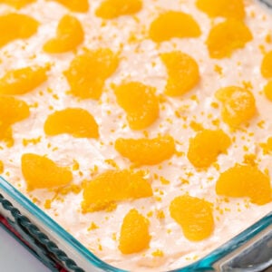 A glass baking dish filled with a creamy dessert topped with evenly spaced mandarin orange segments and orange zest, placed on a red cloth on a white surface.