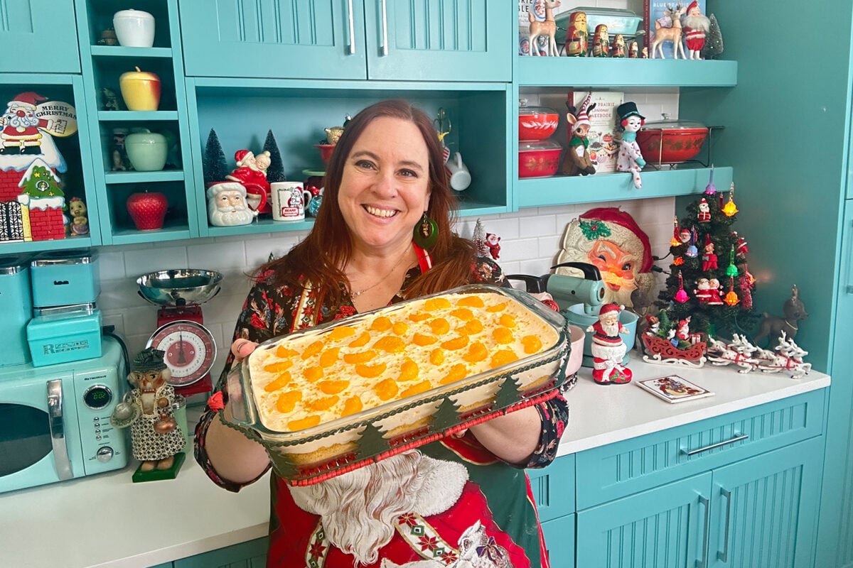 A woman in a festive kitchen holds a casserole topped with sliced mandarins. The kitchen is decorated with Christmas-themed ornaments, including Santa figurines and a small Christmas tree. The woman is smiling and wearing a holiday apron.