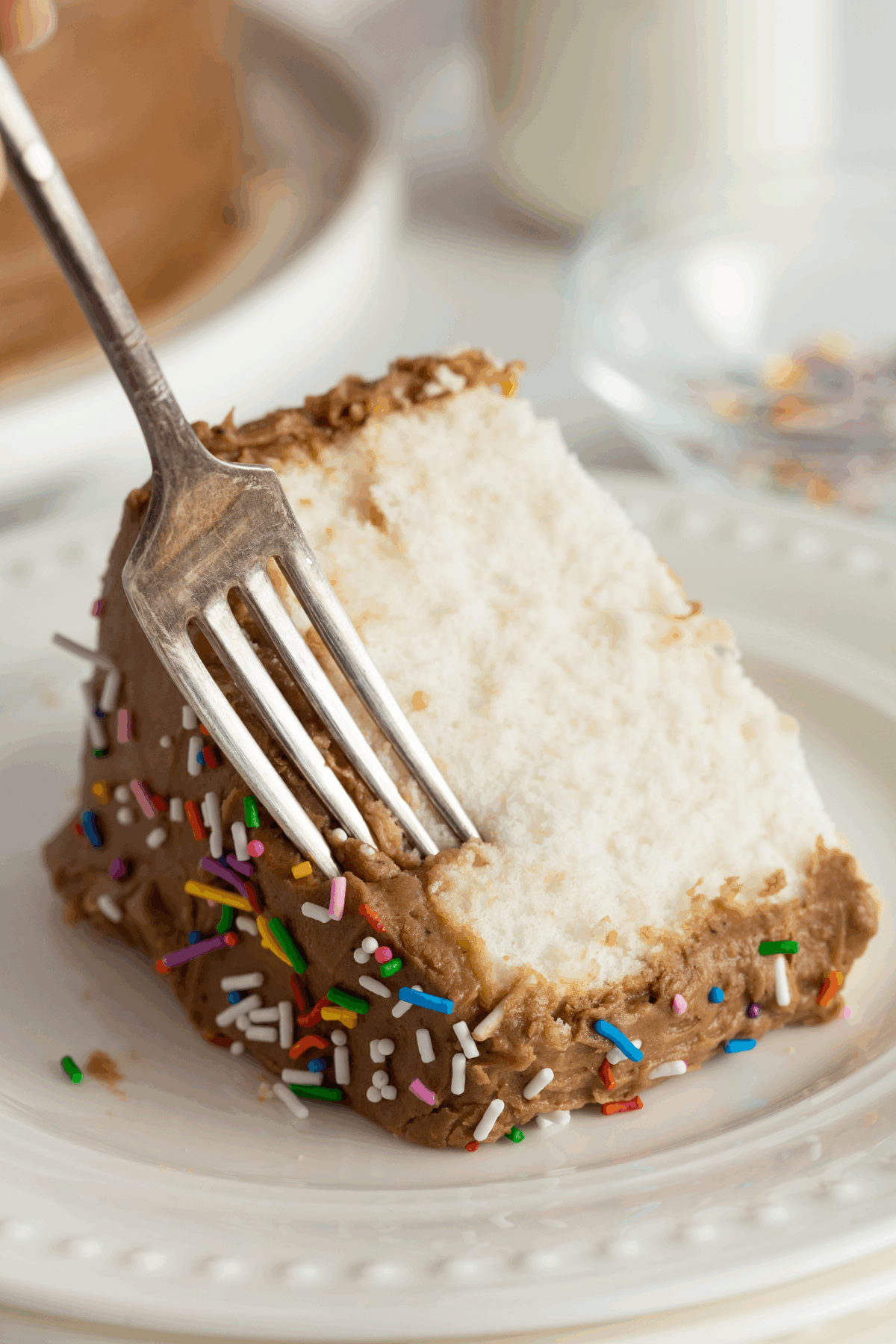 A fork is about to cut into a slice of white cake with brown frosting and colorful sprinkles, served on a white plate. A glass of milk and a bowl of more sprinkles are in the background.