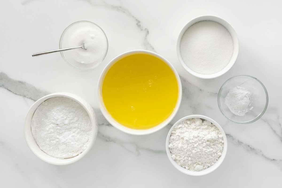 Six bowls on a marble surface contain baking ingredients: melted butter, flour, sugar, powdered sugar, baking powder, and a small bowl of salt with a spoon.