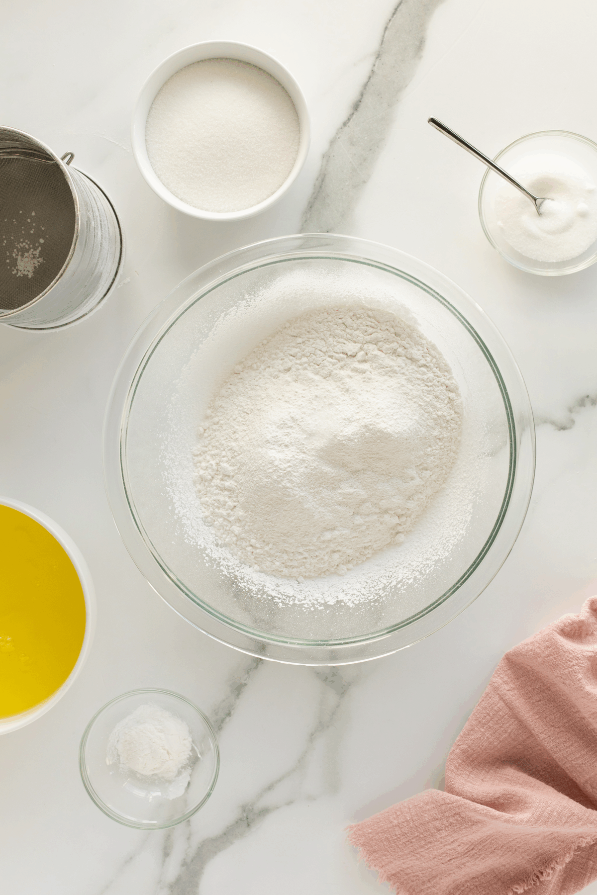 A glass bowl of flour is surrounded by bowls of sugar, baking powder, melted butter, and a sifter on a white marble surface with a pink cloth.