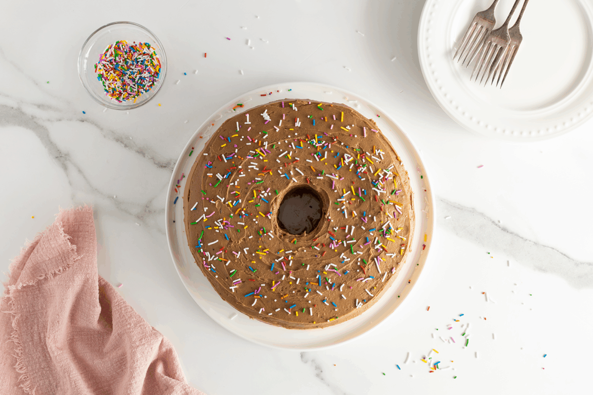 A round cake with brown frosting and colorful sprinkles on a white plate, set on a marble surface. Nearby are a bowl of sprinkles, a pink cloth, and a plate with two forks.