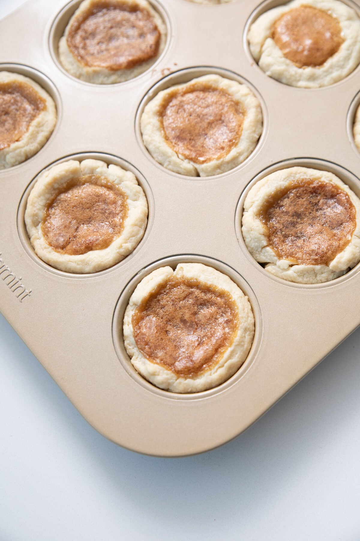 A muffin tin filled with freshly baked butter tarts, each with golden-brown filling and flaky crust edges, on a white surface.