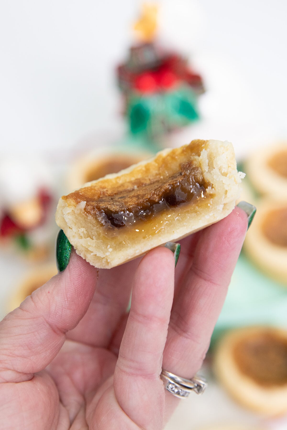 A hand with green nail polish holds half of a butter tart, showing its gooey, caramel-like filling and pastry crust. The background is softly blurred with festive decorations.