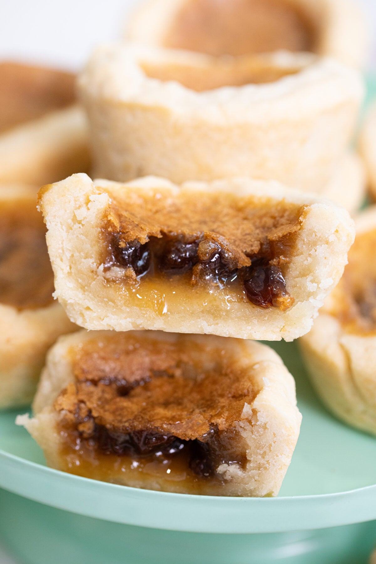 A close-up of stacked butter tarts, with golden flaky crusts and gooey, caramel-like filling visible, including raisins inside the filling. The tarts are on a light green plate.