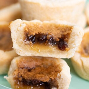 A close-up of stacked butter tarts, with golden flaky crusts and gooey, caramel-like filling visible, including raisins inside the filling. The tarts are on a light green plate.