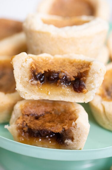 A close-up of stacked butter tarts, with golden flaky crusts and gooey, caramel-like filling visible, including raisins inside the filling. The tarts are on a light green plate.