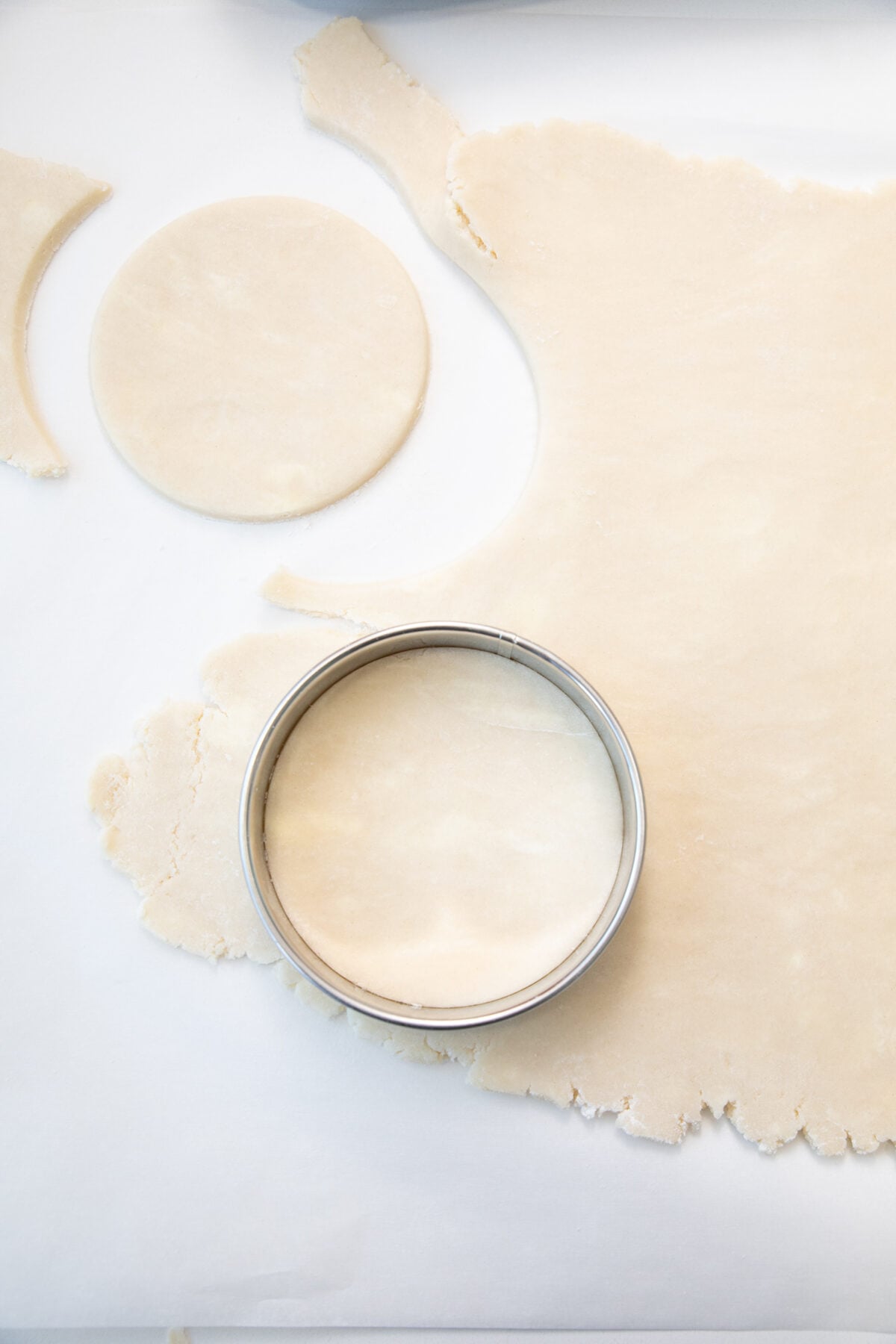 A metal round cutter sits on rolled out dough, with one circular piece cut out and another still inside the cutter, on a white surface.