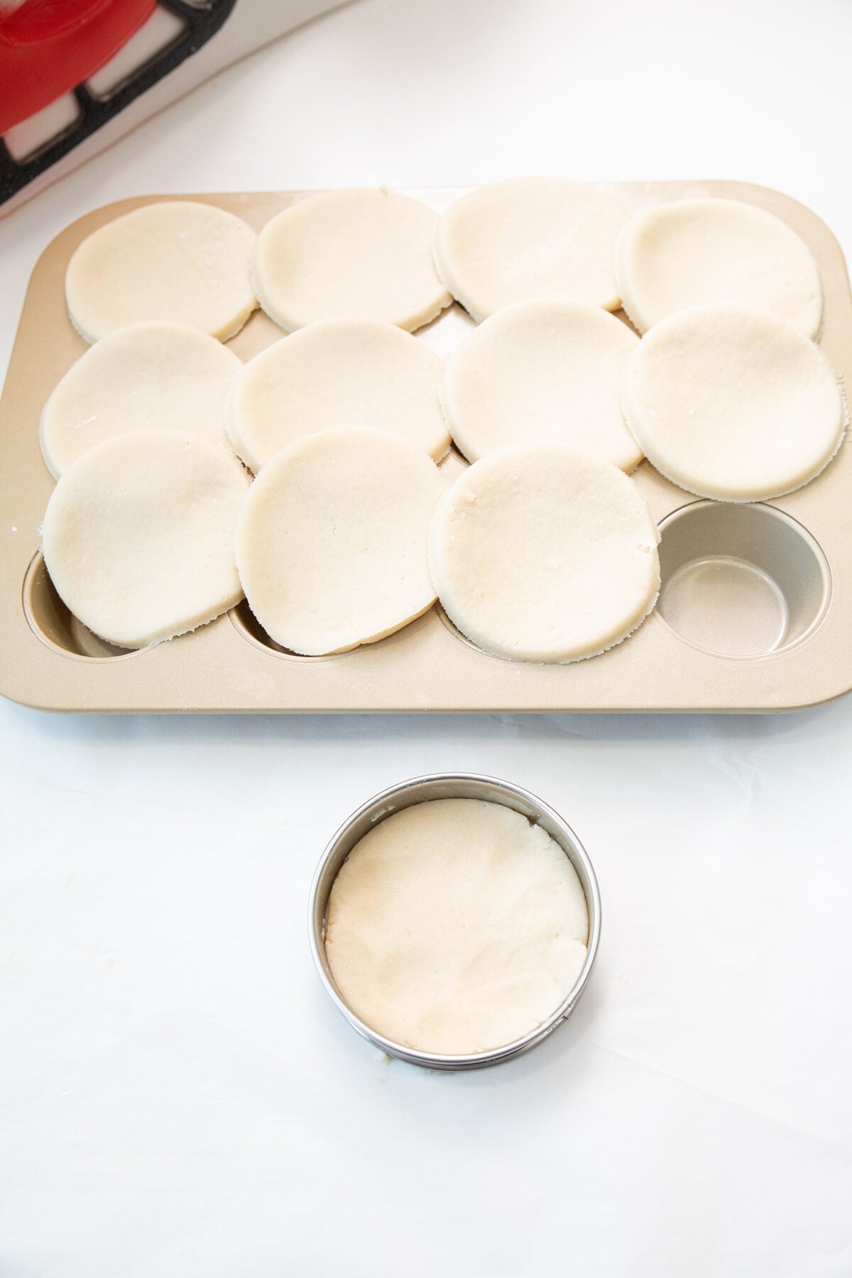 A muffin pan filled with round disks of dough in each cup, with one disk being cut out using a circular metal cutter placed on the white surface nearby.