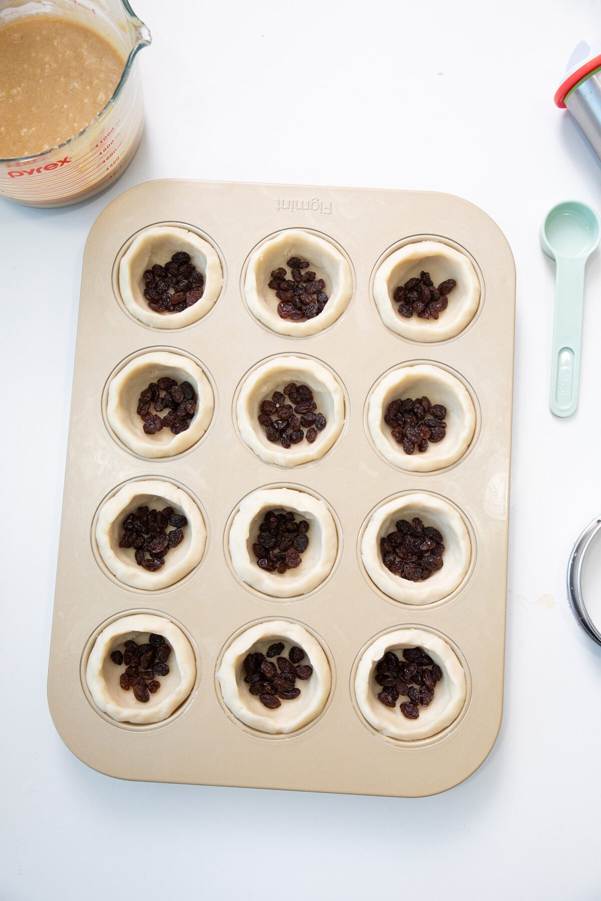 A muffin tin with twelve pastry dough cups, each filled with a small pile of raisins. A measuring cup with batter and a green measuring spoon are nearby on a white surface.