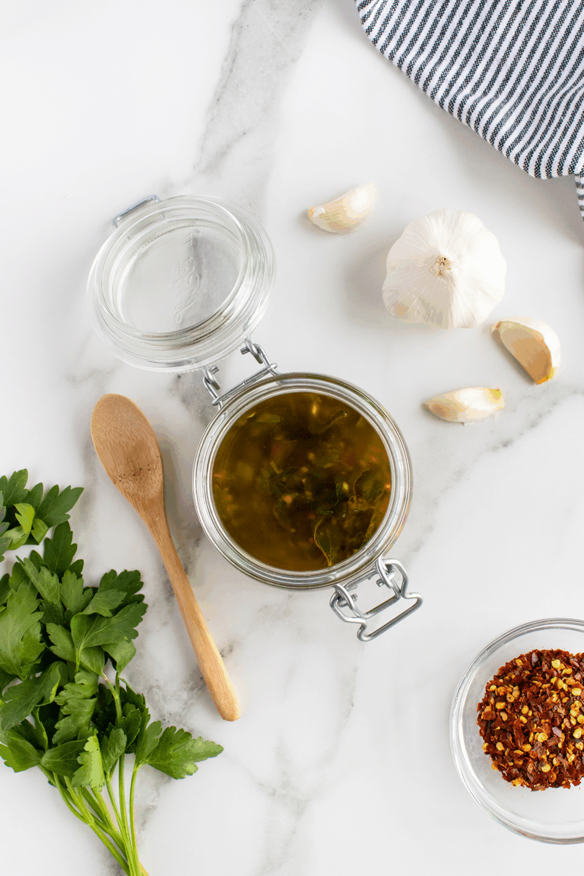 A glass jar of herb-infused olive oil sits open on a marble surface, surrounded by fresh parsley, garlic cloves, a wooden spoon, red pepper flakes in a bowl, and a striped cloth in the corner.