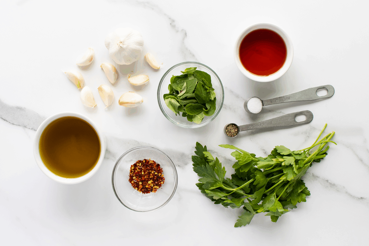 Overhead view of ingredients on a white marble surface: olive oil, garlic cloves, fresh oregano, red pepper flakes, red vinegar, salt, pepper, and a bunch of fresh parsley, with two measuring spoons.