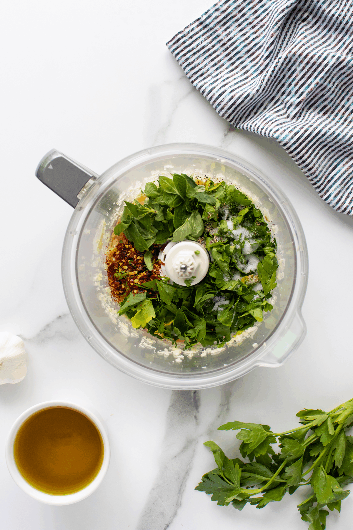 A food processor bowl filled with fresh herbs, spices, and salt sits on a white marble countertop. Nearby are a striped cloth, a bunch of parsley, a cup of olive oil, and a garlic clove.