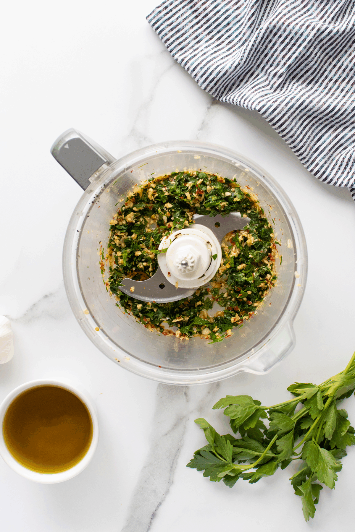 A food processor with chopped herbs and garlic inside sits on a marble counter next to a striped towel, a bunch of fresh parsley, a small bowl of olive oil, and a clove of garlic.