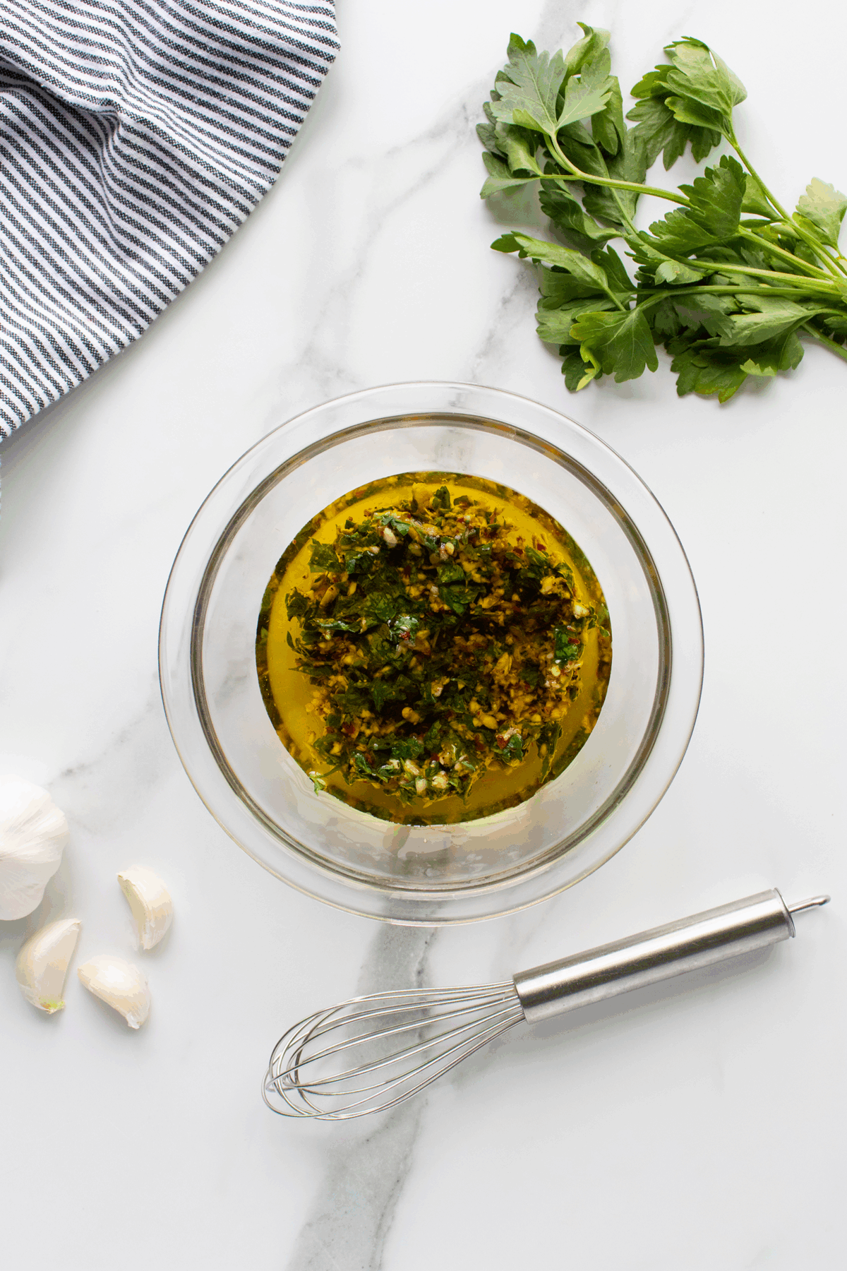 A bowl of herb marinade with chopped parsley and spices sits on a marble surface, surrounded by fresh parsley, garlic cloves, a whisk, and a striped kitchen towel.
