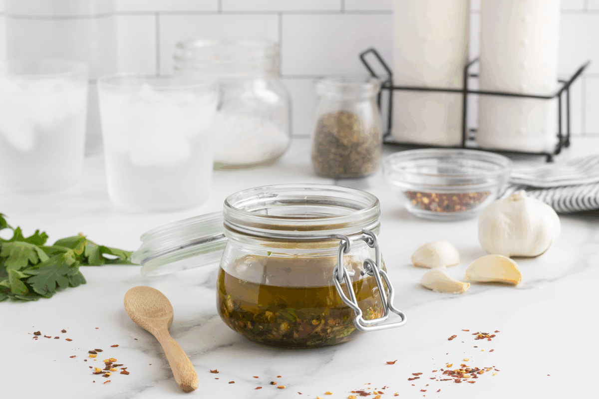 A glass jar filled with homemade oil and herbs sits on a counter next to garlic cloves, parsley, a wooden spoon, and scattered chili flakes. Glasses of water, spices, and candles are in the background.