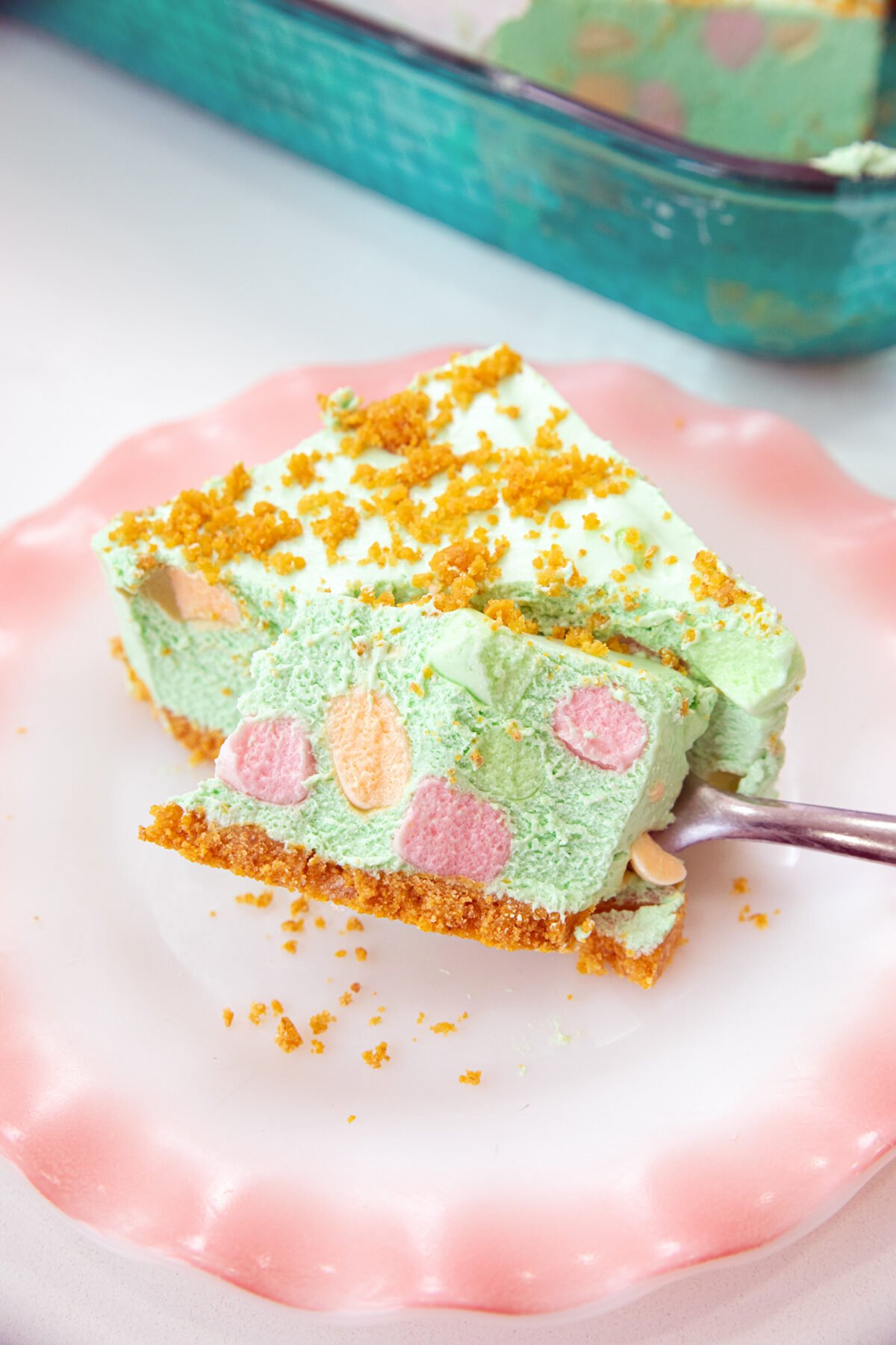 A slice of green, creamy dessert with colorful mini marshmallows and a crumbly graham cracker crust sits on a pink plate, with a fork taking a bite. Crumbs are sprinkled on top.