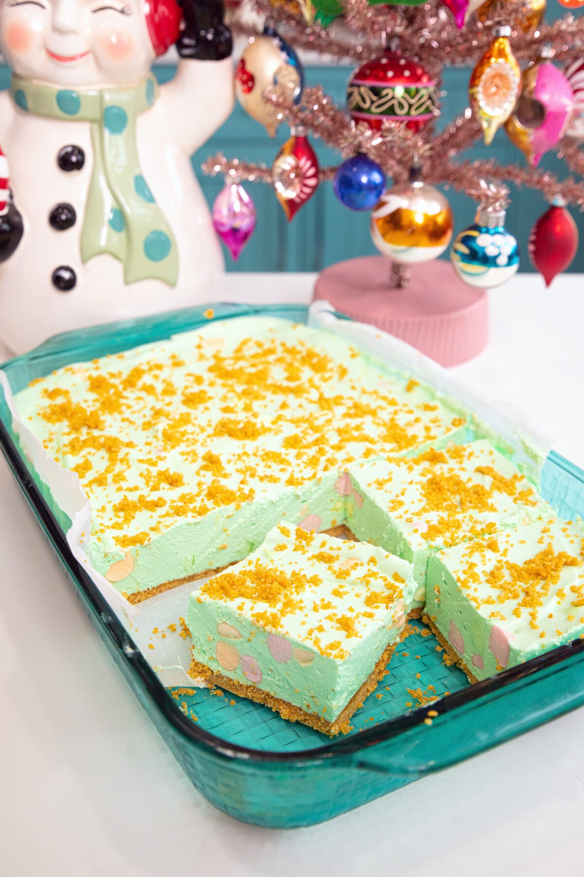 A glass baking dish holds green dessert bars topped with yellow crumbs. Two bars are cut out, revealing a crumbly base. Beside the dish are a vintage ceramic snowman and a pink Christmas tree with colorful ornaments.