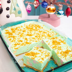 A glass baking dish holds green dessert bars topped with yellow crumbs. Two bars are cut out, revealing a crumbly base. Beside the dish are a vintage ceramic snowman and a pink Christmas tree with colorful ornaments.