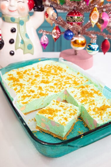 A glass baking dish holds green dessert bars topped with yellow crumbs. Two bars are cut out, revealing a crumbly base. Beside the dish are a vintage ceramic snowman and a pink Christmas tree with colorful ornaments.