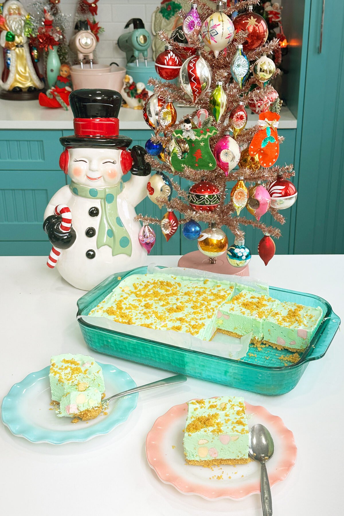 A festive kitchen scene with a vintage snowman cookie jar, a colorful glass ornament Christmas tree, and a dish of green holiday dessert with a graham cracker crust, served on pastel plates with spoons.