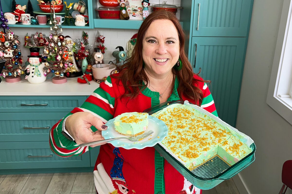 A smiling woman in a festive sweater holds a plate with a slice of green dessert; she stands in a kitchen decorated with colorful Christmas ornaments and a tray of the same dessert.