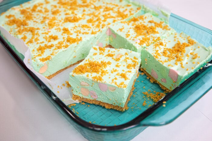 A glass baking dish with squares of light green dessert topped with golden cookie crumbs. The dessert has a crumb crust and colorful marshmallows visible inside the cut pieces.
