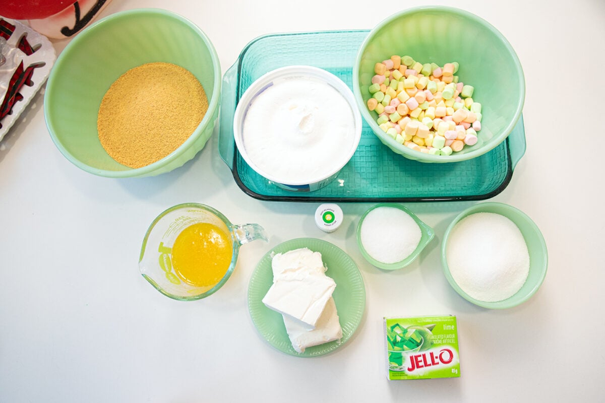 A top-down view of ingredients for a dessert: graham cracker crumbs, whipped topping, pastel mini marshmallows, melted butter, cream cheese, sugar, lime Jell-O mix, and green food coloring on a white surface.