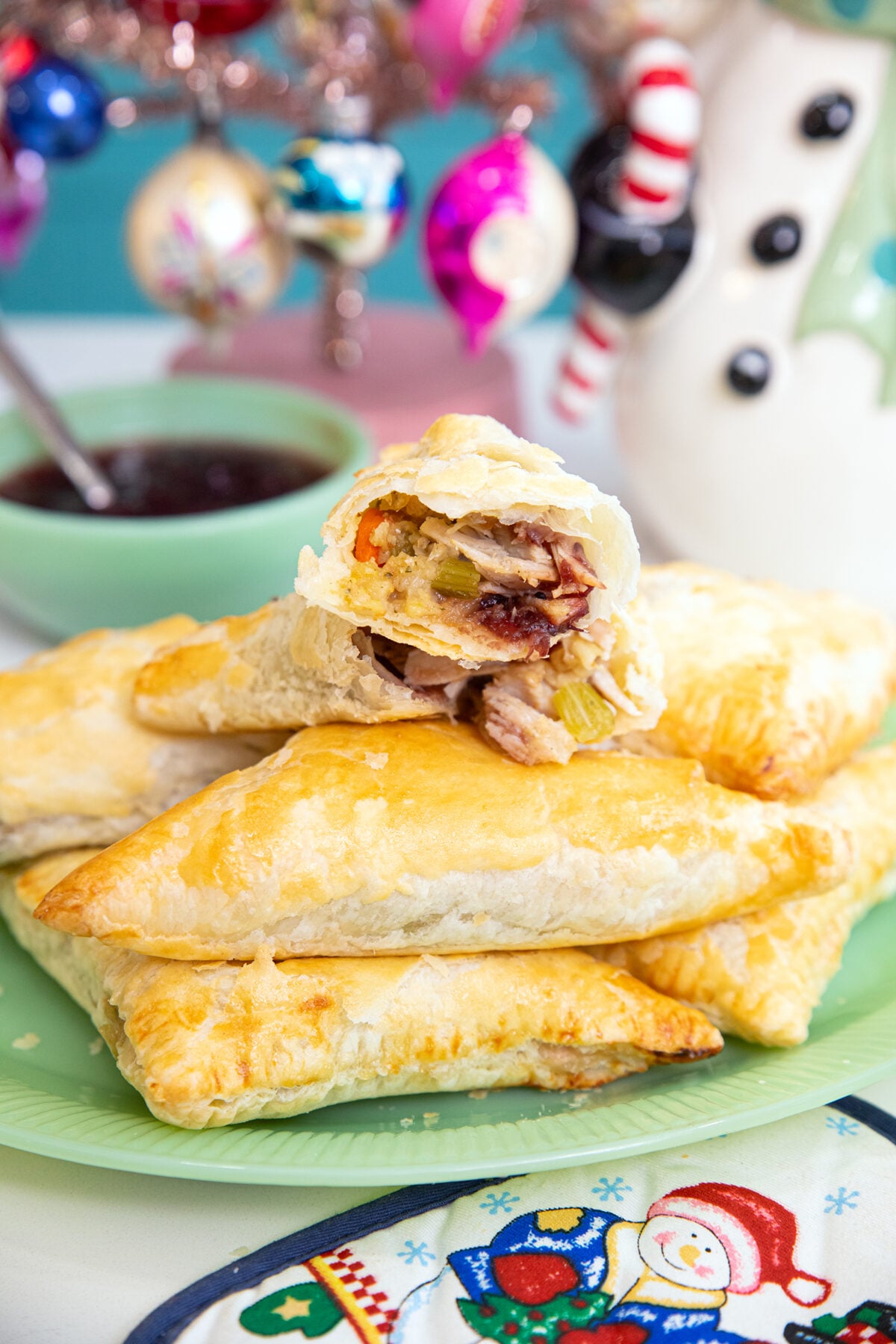 A plate of golden, flaky puff pastry turnovers filled with turkey, vegetables, and cranberry sauce. One turnover is cut open to show the filling. Festive decorations and a bowl of sauce are in the background.