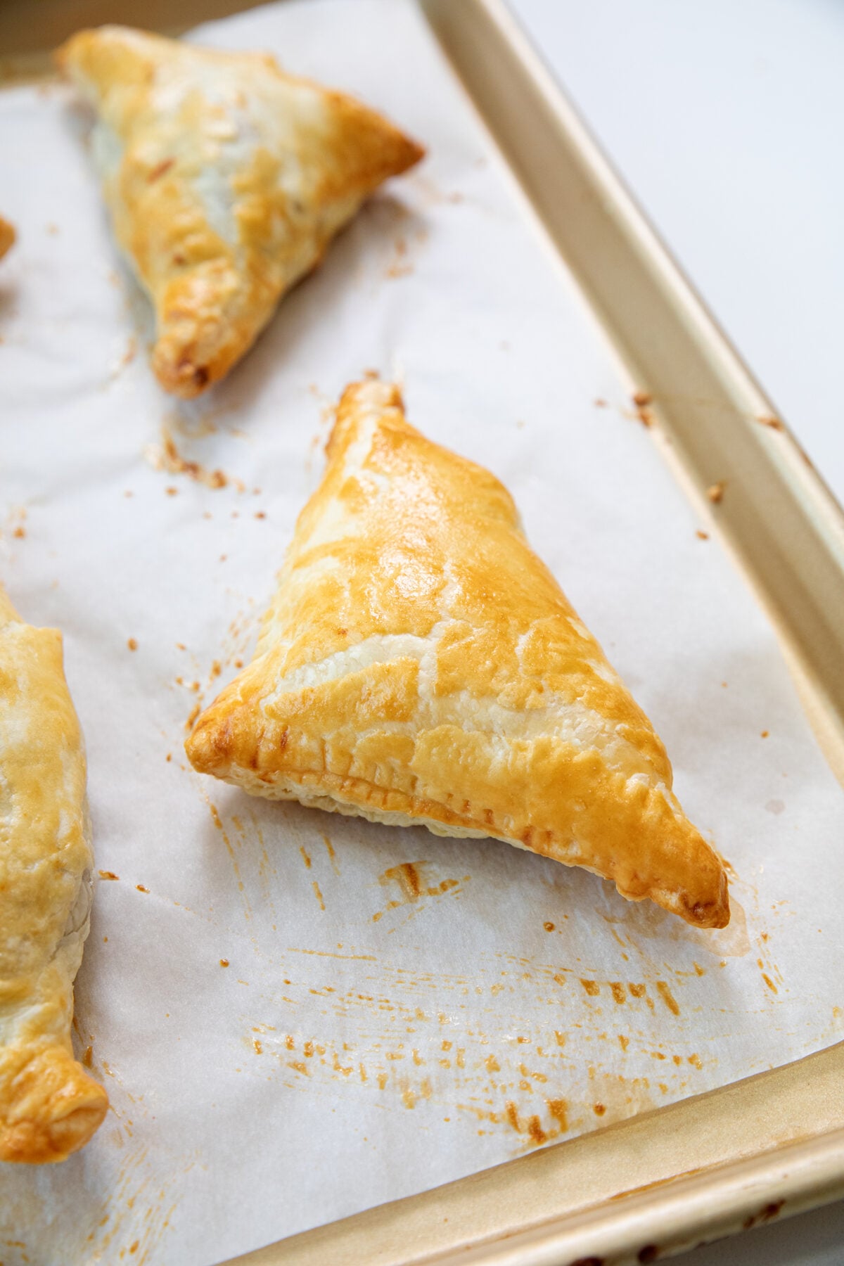 Golden-brown, triangular puff pastries rest on a parchment-lined baking sheet, with flaky layers visible on their surfaces. One pastry is centered in the foreground.
