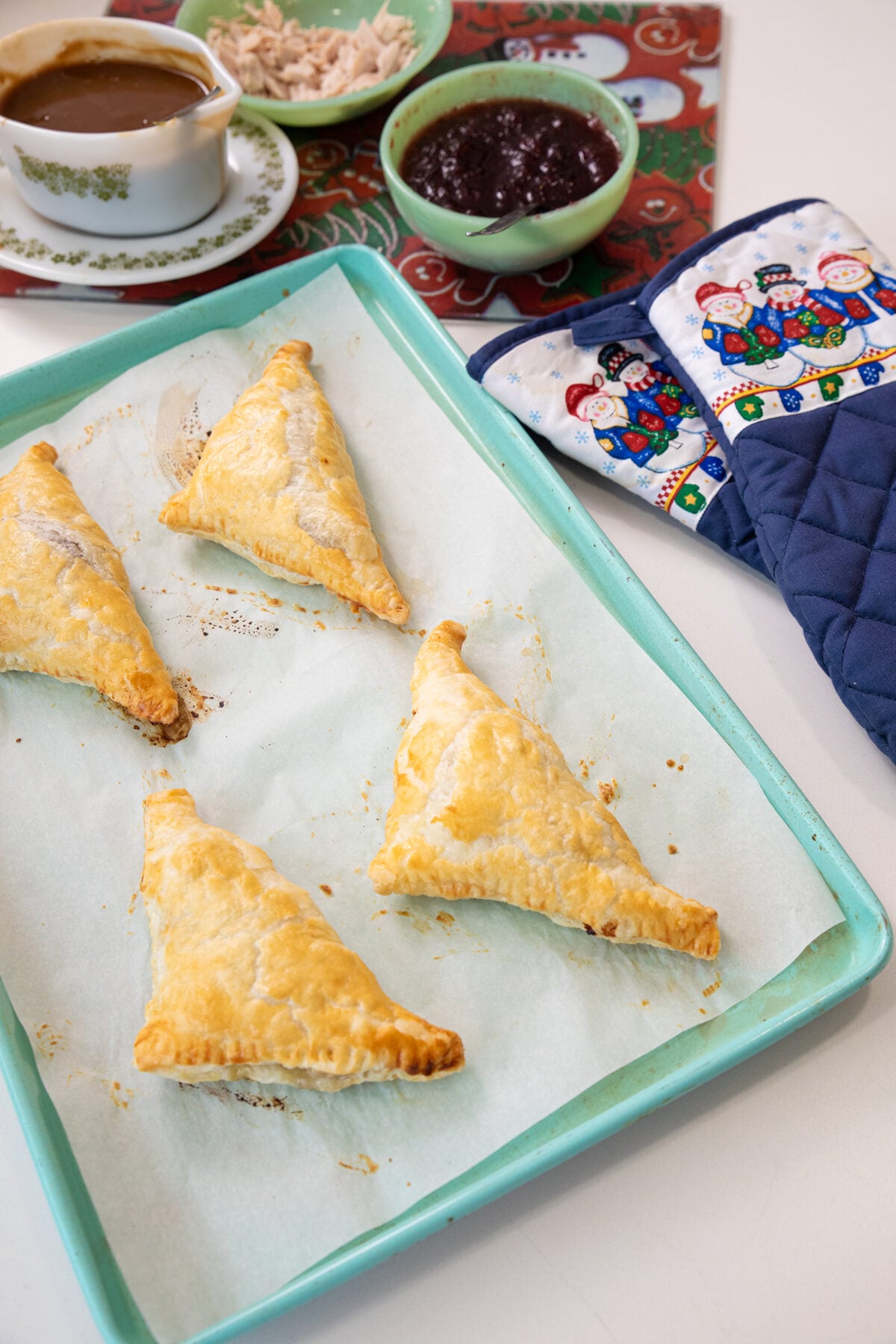 Four golden, flaky puff pastry turnovers on a baking tray lined with parchment paper. In the background are bowls of gravy, cranberry sauce, shredded meat, and a pair of festive oven mitts.