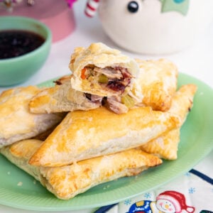 A green plate holds several golden-brown puff pastry turnovers, with one cut open to reveal a savory filling. Nearby are bowls of sauce and jam, and festive holiday decor can be seen in the background.