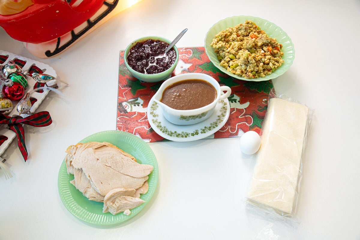 A holiday meal spread includes sliced turkey, stuffing, brown gravy, cranberry sauce, an egg, and a block of dough on a white table with festive decorations and a red and white placemat.