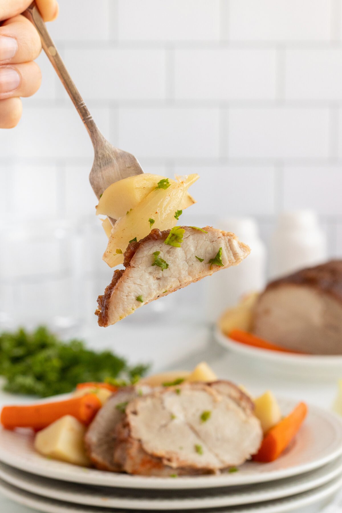 A fork holds a bite of roasted pork and sliced potatoes garnished with herbs, with a plate of sliced pork, potatoes, and carrots on a table in the background.