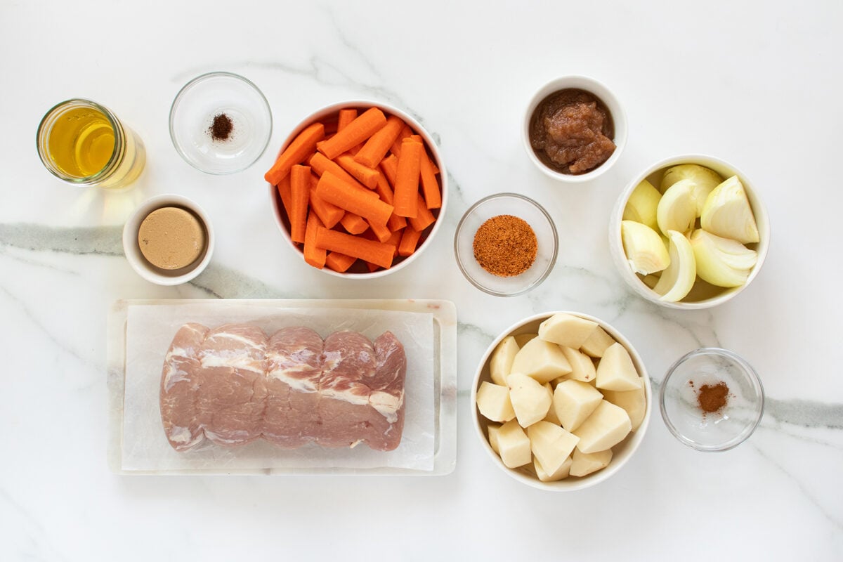 A top-down view of ingredients on a white surface: raw pork, chopped potatoes, carrot sticks, onion wedges, spices, apple sauce, mustard, oil, and brown paste in separate bowls.