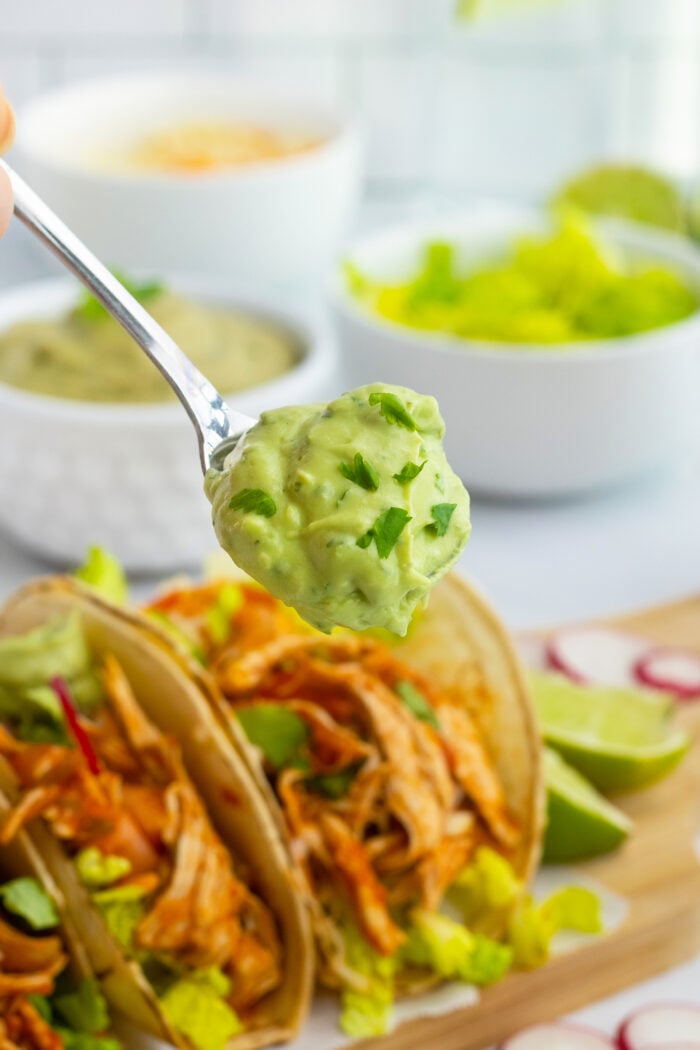 A spoonful of creamy guacamole is held above a plate with chicken tacos, surrounded by bowls of lettuce, green sauce, and lime wedges in the background.